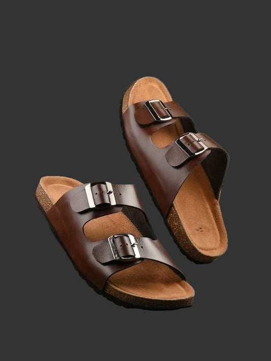 Brown sandals with buckles on a wooden surface with a plant in the background